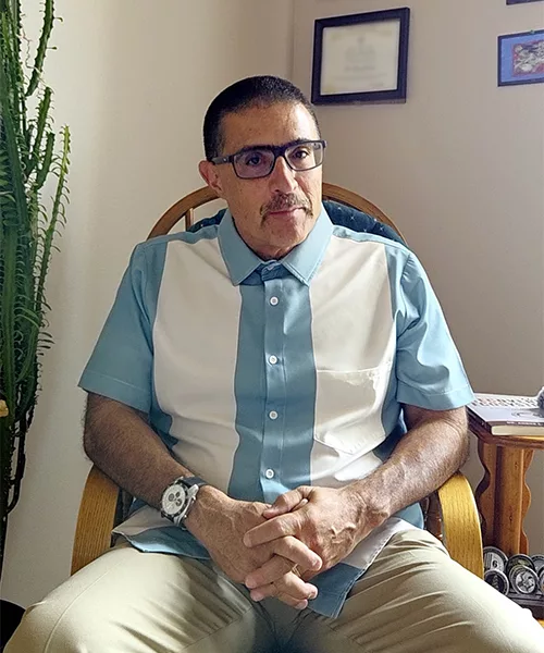 A man sitting up straight in his char with his hands clasp looking at the camera