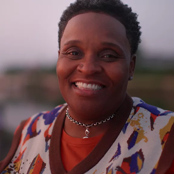 A close up of Mona smiling at the camera outdoors wearing a colorful sweater vest